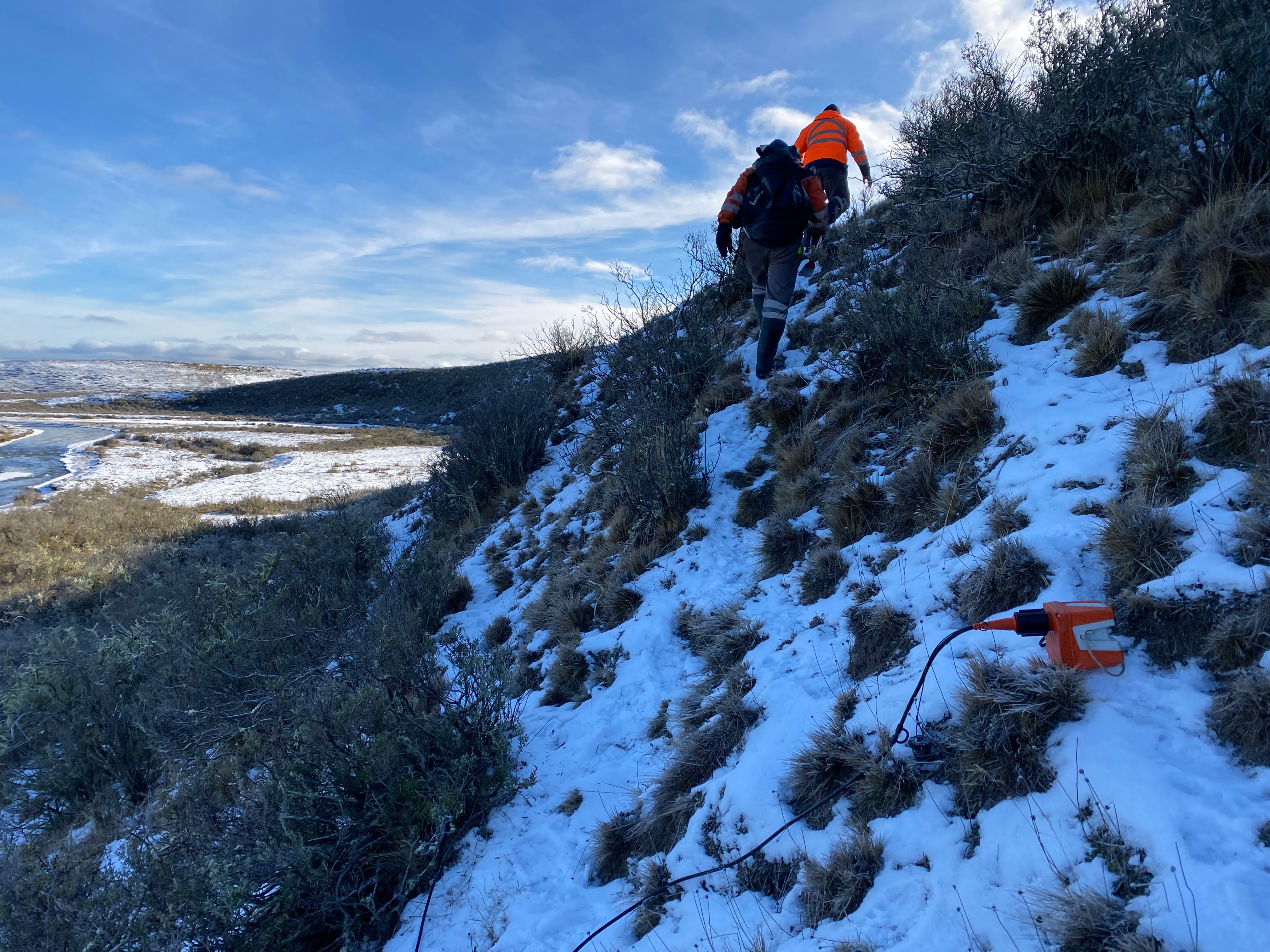 Sensor sísmico naranja sobre nieve con técnico cruzando un lago helado